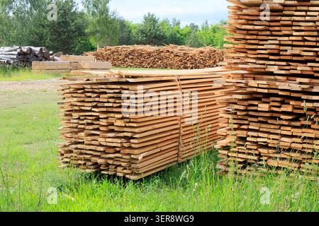 Sawn timber boards stacks at an open air lumberyard. Packed wooden planks piles sit on green grass field near logs stack at sawmill factory under clou Stock Photo