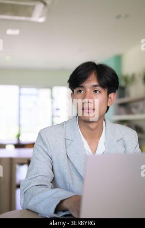 Authentic vertical lifestyle portrait of a young non-binary Thai Isan professional or student working on a computer and looking away thoughtfully. Ser Stock Photo