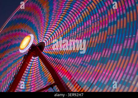 Long exposure digital capture of a ferris wheel turning at night at the state fair, creating vivid circular light trails in red, pink, blue, cyan, ora Stock Photo