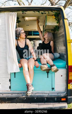 Two women relax in a cozy camper van with turquoise storage, chatting and stretching under the open rear doors, enjoying a sunny holiday road trip in Stock Photo