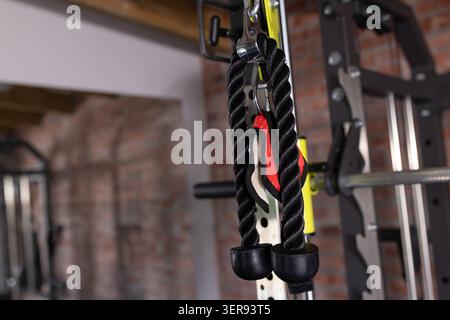 Black twisted tricep rope is hanging from metal carabiner on yellow support, with red strap visible Stock Photo