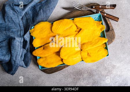 A vibrant display of dried mango slices elegantly arranged on a turquoise ceramic plate. Fork and knife rest nearby, set on a rustic wooden board over Stock Photo