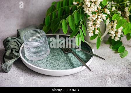 Minimal table setting with a white plate, textured water glass, cutlery and linen, styled with fresh acacia flower and green leaves against a soft neu Stock Photo