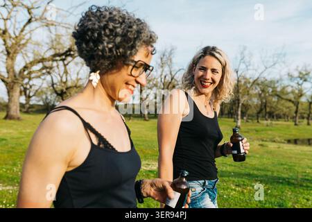 Two women friends laugh together while walking in a sunny park, holding cold drinks and enjoying a relaxed spring holiday getaway in nature with warm Stock Photo