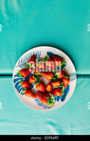 Top view of ripe strawberries on a white plate with blue tropical leaf pattern, placed on a turquoise fabric background. Bright summer food concept. Stock Photo