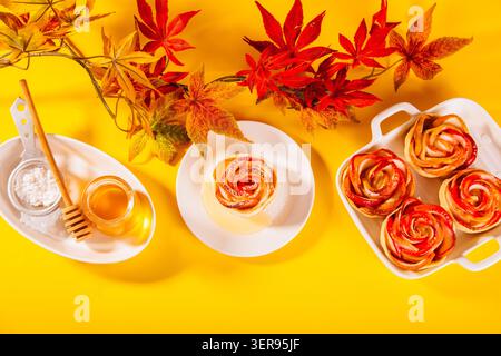 Delicate rose-shaped baked apple desserts arranged artfully on a white plate, surrounded by autumn leaves, powdered sugar, and a jar of honey for a pe Stock Photo