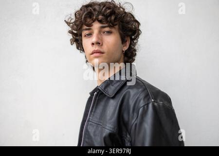 Confident curly haired man posing in a black leather jacket against a light wall, modern lifestyle portrait with relaxed attitude, minimal background Stock Photo