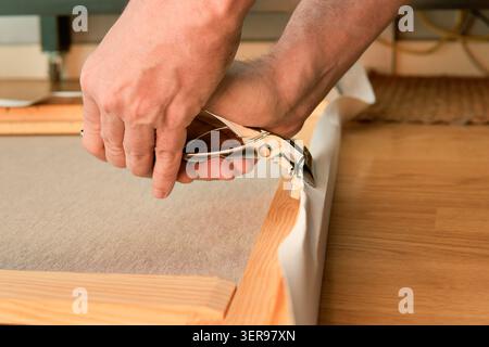 Close-up of hands stretching canvas fabric over a wooden stretcher frame, using a metal stapler to fasten the edge with wooden staple marks in a works Stock Photo