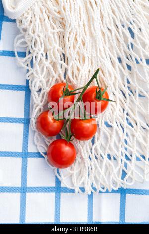 Close-up of ripe cherry tomatoes on the vine resting on a white mesh bag over a blue checkered tablecloth, creating a bright, fresh and minimal food b Stock Photo