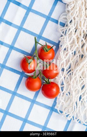Flat lay of fresh cherry tomatoes on a blue and white checkered kitchen cloth beside a reusable cotton net bag, creating a clean, minimal concept for Stock Photo