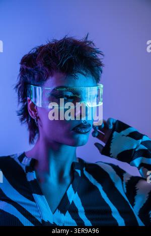 Close-up portrait of a young woman in neon blue and purple light, wearing a futuristic virtual reality visor and cyberpunk style makeup, posing in a s Stock Photo