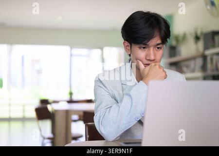 Authentic horizontal lifestyle portrait of a young non-binary Thai Isan professional or student looking down at a computer screen with a hand on chin. Stock Photo