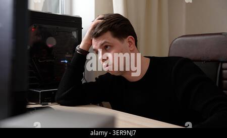 Young man in black sweater sitting at desk looking at computer screen with worried expression, hand on head, showing stress and frustration while work Stock Photo