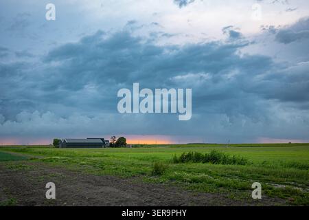Dark storm clouds sweep across a rural farm field at sunset, a dramatic blue-gray sky above a green field and distant barn Stock Photo