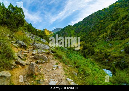 Dirt and stone hiking trail winds up a rocky slope covered in vibrant green vegetation in Kyrgyzstan. A natural stream flows alongside, enhancing the Stock Photo