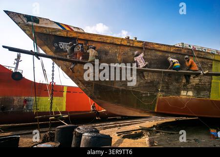 Dhaka, Bangladesh - 03 June 2026: View of workers sitting on a wooden scaffolding while painting and repairing the hull of a large ship in a shipyard Stock Photo