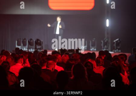 Conference speaker on stage presenting to attentive audience in dim auditorium with red stage lighting, professional event atmosphere, public speaking and live presentation. Stock Photo