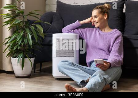 smiling woman sitting on the floor by air purifier at home. air quality and microclimate Stock Photo