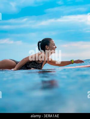 Young Woman Focused Paddling On Surfboard In Calm Sea, Determined Athletic Training With Concentrated Posture Stock Photo