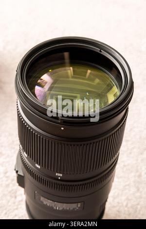 Close-up view of a telephoto camera lens resting on a light-colored textured surface. Lens features focal length markings and reflections of an indoor Stock Photo