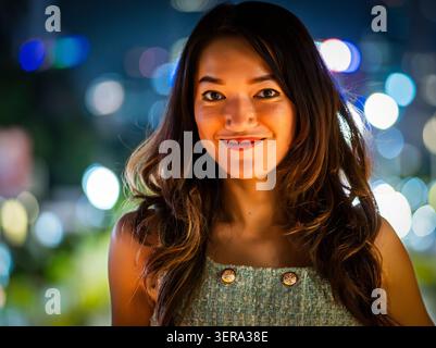 Smiling young woman with long dark hair in a sleeveless top, softly lit at night with colorful city lights blurred in the background. Stock Photo