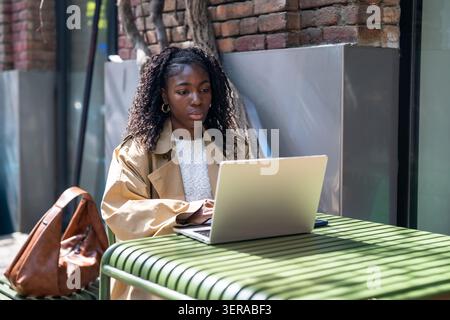 Thoughtful african american woman student working on laptop outdoors in city cafe on sunny day. Stock Photo