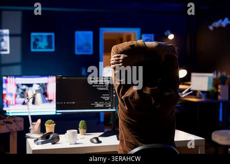Programmer doing software quality assurance, stretching arms behind back after reading source code, feeling fatigued. IT expert taking break from work to relax muscles after inspecting coding. Stock Photo