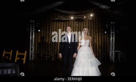 Elegant young couple, man in black tuxedo, woman in white tiered gown, walk hand in hand. Dramatic lighting highlights their confident pose against da Stock Photo