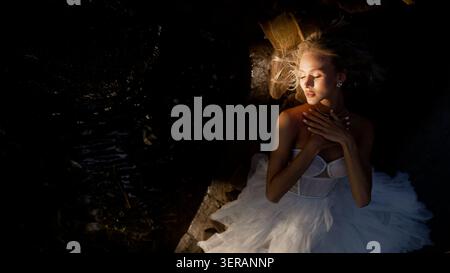 Young woman in white elegant dress lies peacefully on dark rocks. Golden hour light illuminates her face, hair, shoulders, creating dramatic chiaroscu Stock Photo