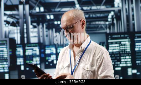 Aged professional navigating network of server rigs in industrial headquarters, with iT programmer with tablet inspecting and optimizations in high tech data center, preventing system concerns. Stock Photo