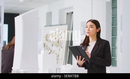 In property management office, contractor uses tablet to evaluate and estimate variables as they work with 3D printed architectural prototype, engineer working to design urban area with housing. Stock Photo