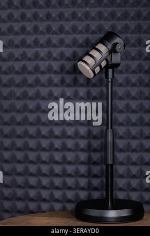 Black dynamic microphone mounted on an adjustable desktop stand, placed on a wooden surface against a dark grey acoustic foam panel background, ideal Stock Photo