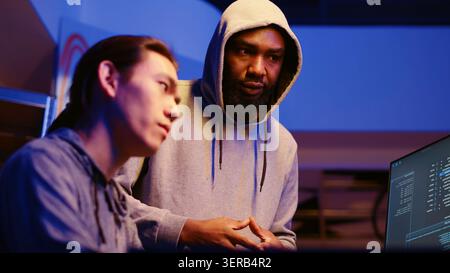 BIPOC hackers in dimly lit room discussing ways to breach defenses and exploit weaknesses in PC systems. Stock Photo