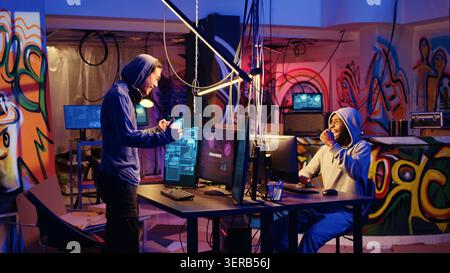 Hackers in abandoned warehouse celebrating after successfully using phishing technique that tricks users into revealing sensitive information, happy cybercriminals excited after stealing passwords. Stock Photo