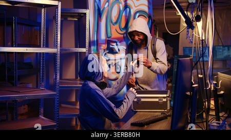 African American hacker in secret underground base excited to show colleague SSD with finished spyware virus they will use to gather data from users computers without their knowledge. Stock Photo
