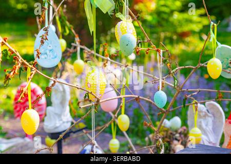 Colorful Easter eggs hanging on tree branches in a sunny garden with blurred plants and small statues background Stock Photo