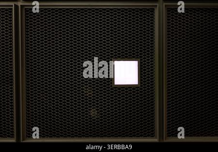 Minimalist industrial background featuring dark metal mesh wire panels with a single glowing white square LED light fixture in center Stock Photo