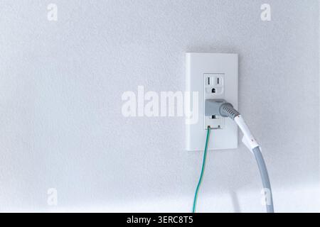A white electrical outlet with two grey power cables plugged in, with exposed wire ends visible on a white wall. The in-progress electrical work detail documents renovation and home construction work. Stock Photo