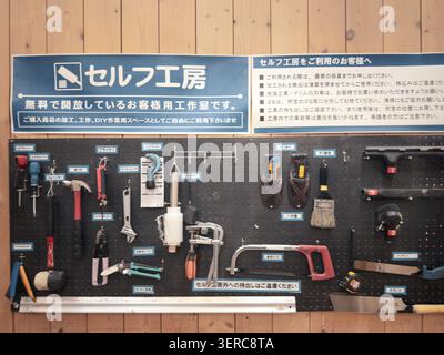 Hand tools including hammers, saws, and clamps are arranged on a black pegboard wall in a Japanese self-service DIY workshop. A sign above displays the facility's free-use policy in Japanese. Stock Photo
