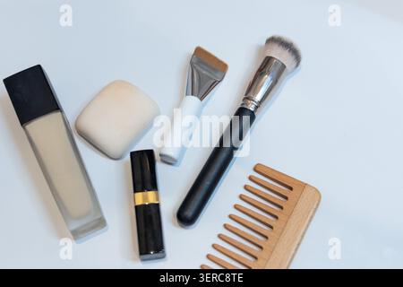 A flat lay overhead shot of makeup items - foundation bottle, makeup sponge, lipstick, contour brush, powder brush, and wooden comb - arranged diagonally on a white surface. Stock Photo