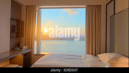 Luxury hotel bedroom interior. Large window view of golden sunrise over city river. Modern furniture, white bedding, warm morning light. High-end trav Stock Photo