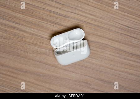 White charging case rests open on wooden surface. Clean, minimalist design with soft lighting. Earbuds not visible inside the case. Wood grain texture Stock Photo