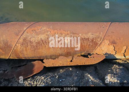 A close-up focus on a metal pipeline at the water's edge. The surface is covered with thick reddish-brown rust, with large areas of coating peeling of Stock Photo