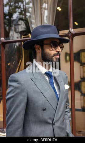 A stylish, bearded man wearing a suit, a classic blue fedora hat, sunglasses, stands outside an elegant shop window in London. Stock Photo