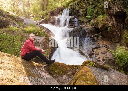 Man in a red jacket sitting on rocks observing a forest waterfall Stock Photo