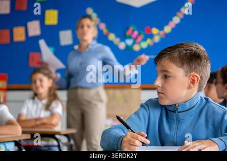 Primary school student sitting at his desk and paying attention during a classroom lesson while a teacher explains concepts in the background. Stock Photo