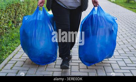 Person carrying two blue garbage bags for waste disposal and recycling on sidewalk Stock Photo