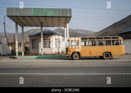 An old yellow Soviet-era bus with passengers visible inside is parked next to a derelict roadside gas station in a rural, mountainous region of Armeni Stock Photo