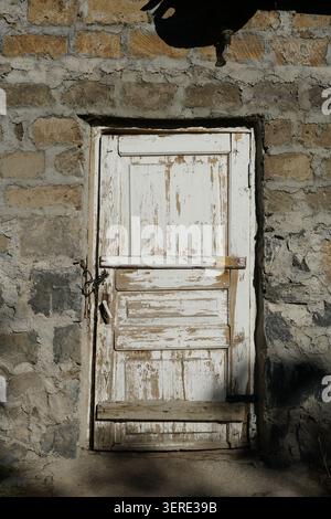 A close-up view of an aged white wooden door with extensively peeling paint, set within a rustic stone wall. The door features a simple latch and chai Stock Photo