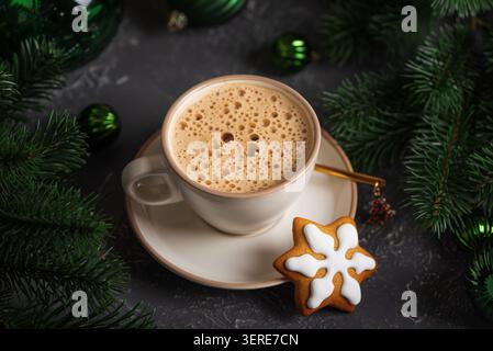 Mug of frothy coffee with a festive gingerbread snowflake cookie surrounded by green christmas tree branches and ornamental baubles on a dark backgrou Stock Photo
